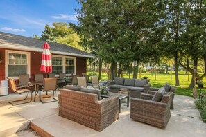 A patio with wicker furniture, a red and white umbrella, and a view of a green lawn and trees. - Well-appointed 3-bedroom house, sleeps 8 with gorgeous sunsets! (Manistee)