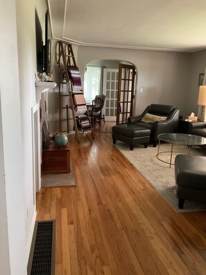 A living room with a wooden floor, a black leather sofa, a glass coffee table, and a staircase with a wooden railing. - Well-appointed 3-bedroom house, sleeps 8 with gorgeous sunsets! (Manistee)