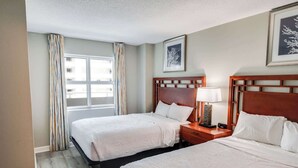 A hotel room with two beds, a window with curtains, a bedside table with a lamp, and framed artwork on the wall. - Amazing Oceanfront View Unit with Pool Access (Myrtle Beach)