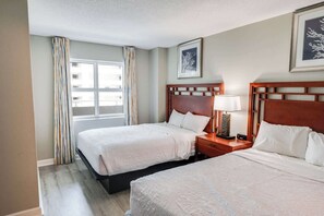 A hotel room with two beds, a window with curtains, a bedside table with a lamp, and framed artwork on the wall. - Amazing Oceanfront View Unit with Pool Access (Myrtle Beach)