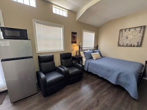 A compact bedroom with a bed, two chairs, a refrigerator, and a lamp.