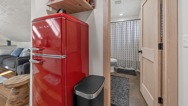 Ein kompakter Wohnraum mit einem roten Kühlschrank, einer Mikrowelle, einer kleinen Mülltonne, einem grauen Sofa und einer Holztür, die zu einem Badezimmer führt.