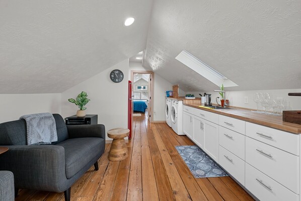 A compact living space with a sofa, a small table, a clock, a washing machine, and a kitchen area with cabinets and a countertop. - Luxurious Furnished Loft Suite - GREAT for Traveling Nurses/Students (Sioux Falls)