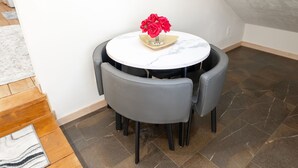 Ein runder Tisch mit einer Vase roter Blumen, umgeben von vier grauen Stühlen in einer Ecke eines Raumes mit Fliesenboden und einer Holztreppe.