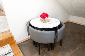 Ein runder Tisch mit einer Vase roter Blumen, umgeben von vier grauen Stühlen in einer Ecke eines Raumes mit Fliesenboden und einer Holztreppe.