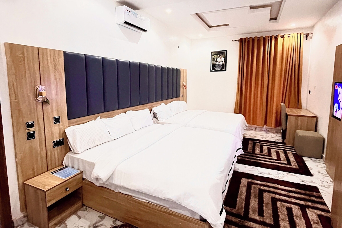 A hotel room with a large bed, wooden headboard, a small bedside table, a wall-mounted air conditioner, a brown rug, and a television on the wall.