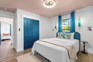 Ein Schlafzimmer mit einem großen Bett, einer blauen Tür, einem Fenster mit Vorhängen und einem kleinen Tisch.