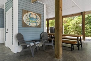 A covered porch with Adirondack chairs and a wooden table, a wooden wall with a painted crab, and a view of greenery. - The Crabby Buffalo𓇼Golf Cart+Outdoor Oasis+Cozy (Oak Island)