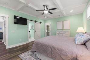 A bedroom with a bed, a ceiling fan, a TV, and a door leading to a bathroom. - The Crabby Buffalo𓇼Golf Cart+Outdoor Oasis+Cozy (Oak Island)