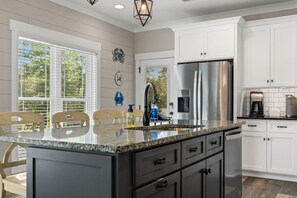 A kitchen with a central island, white cabinets, stainless steel appliances, and a window with blinds. - The Crabby Buffalo𓇼Golf Cart+Outdoor Oasis+Cozy (Oak Island)