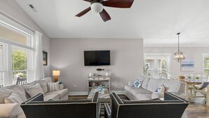 A living room with a ceiling fan, a flat-screen TV, a dining area with a table and chairs, and a bookshelf. - The Crabby Buffalo𓇼Golf Cart+Outdoor Oasis+Cozy (Oak Island)