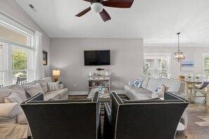 A living room with a ceiling fan, a flat-screen TV, a dining area with a table and chairs, and a bookshelf. - The Crabby Buffalo𓇼Golf Cart+Outdoor Oasis+Cozy (Oak Island)
