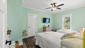 A bedroom with two beds, a ceiling fan, a television, and a window with blinds. - The Crabby Buffalo𓇼Golf Cart+Outdoor Oasis+Cozy (Oak Island)