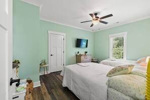 A bedroom with two beds, a ceiling fan, a television, and a window with blinds. - The Crabby Buffalo𓇼Golf Cart+Outdoor Oasis+Cozy (Oak Island)