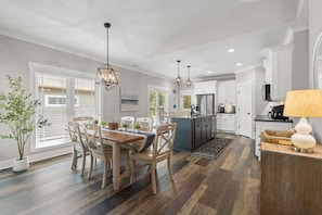 A modern kitchen with a dining area, featuring a central island, pendant lighting, and a view of the outdoors. - The Crabby Buffalo𓇼Golf Cart+Outdoor Oasis+Cozy (Oak Island)