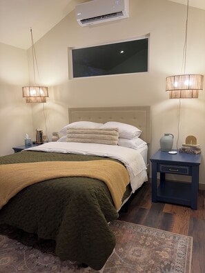 A bedroom with a bed, bedside tables, a nightstand, a window, and an air conditioning unit.