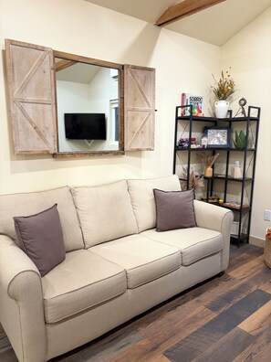 A beige sofa with two cushions, a wooden-framed mirror, a black shelving unit with various items, and a television on the wall.