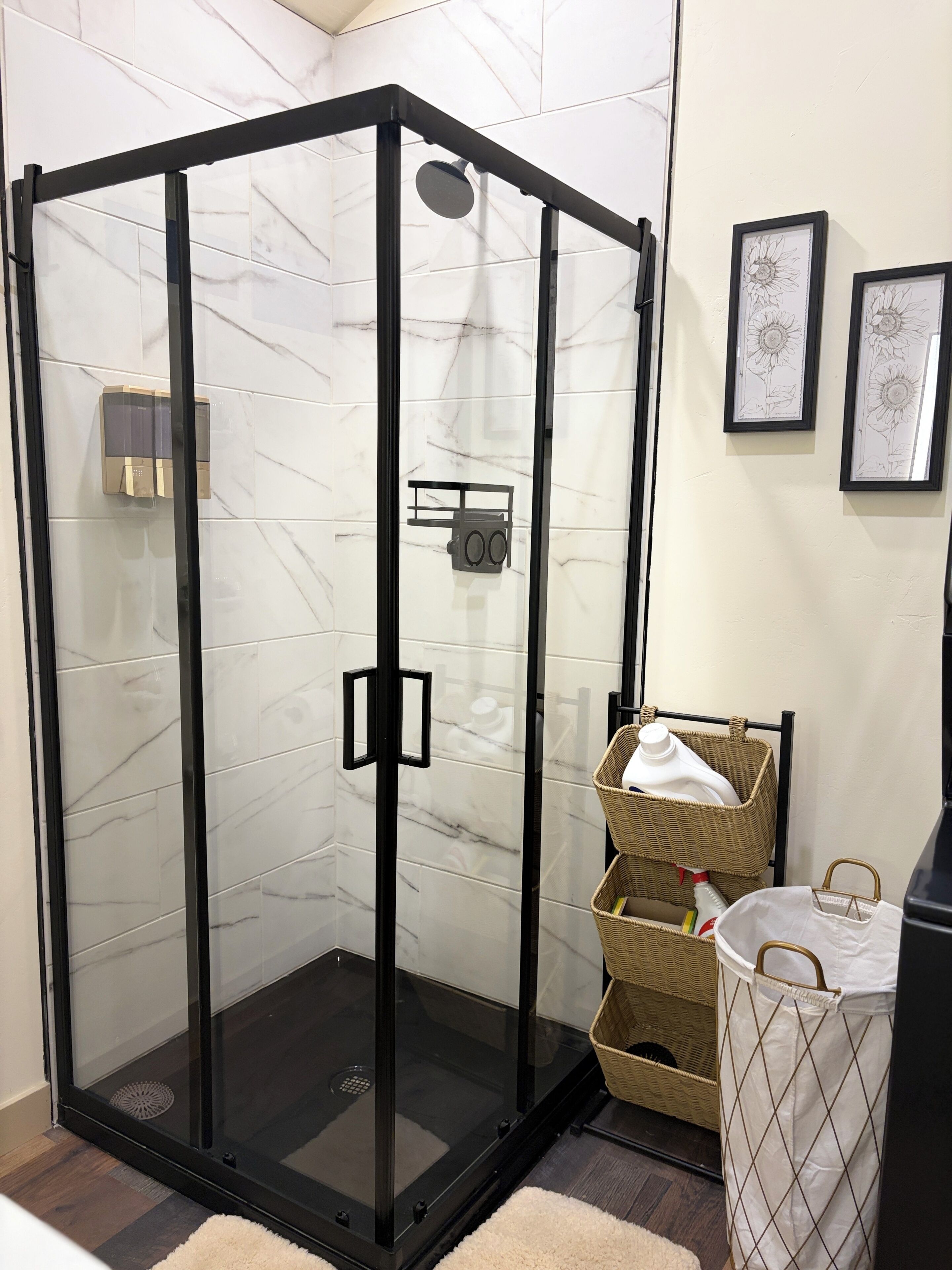 A glass-enclosed shower with a control panel, a wicker basket with a white bag, and a trash bin.
