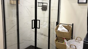 A glass-enclosed shower with a control panel, a wicker basket with a white bag, and a trash bin.