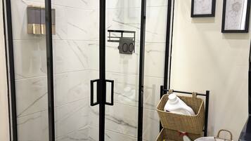 A glass-enclosed shower with a control panel, a wicker basket with a white bag, and a trash bin.