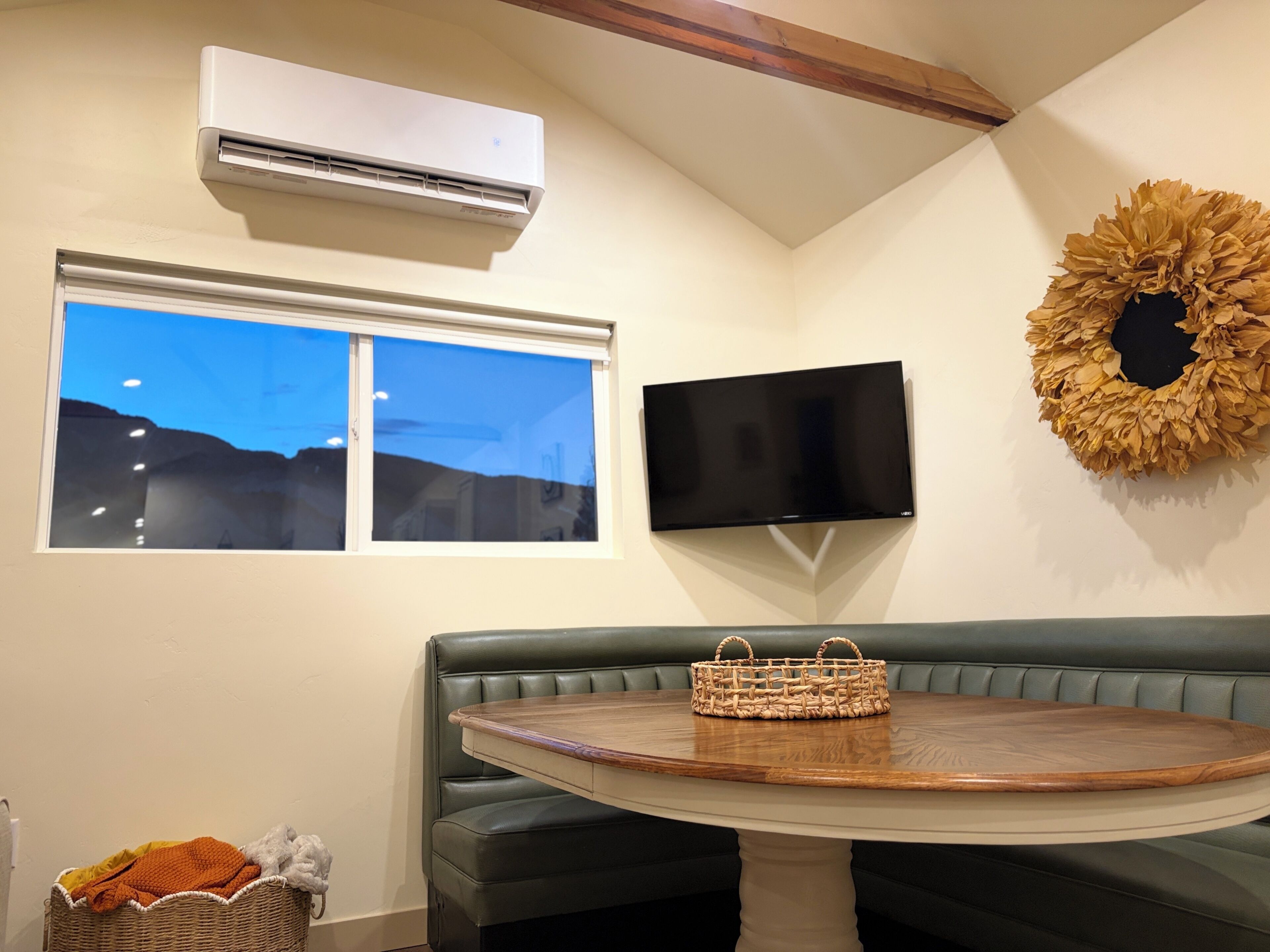 A room with a green leather bench, a round wooden table, a woven basket, a flat-screen TV, and an air conditioning unit.