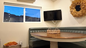 A room with a green leather bench, a round wooden table, a woven basket, a flat-screen TV, and an air conditioning unit.