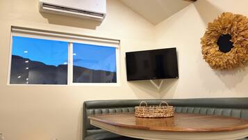 A room with a green leather bench, a round wooden table, a woven basket, a flat-screen TV, and an air conditioning unit.