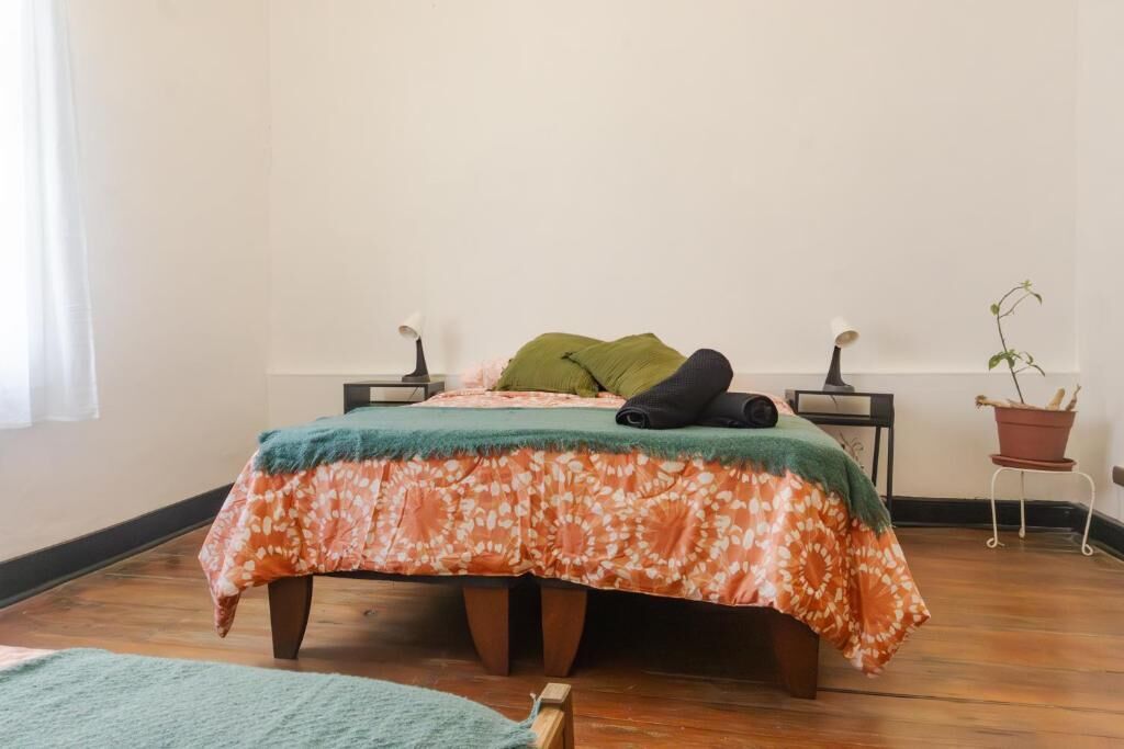 Un dormitorio con cama, mesita de noche, planta en maceta y ventana con cortinas.