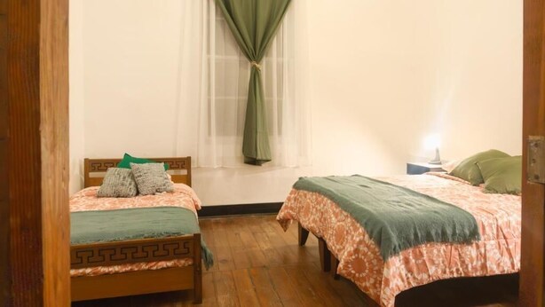 Habitació amb dos llits, una finestra amb cortines fines, una tauleta de nit amb una làmpada i un terra de fusta.
