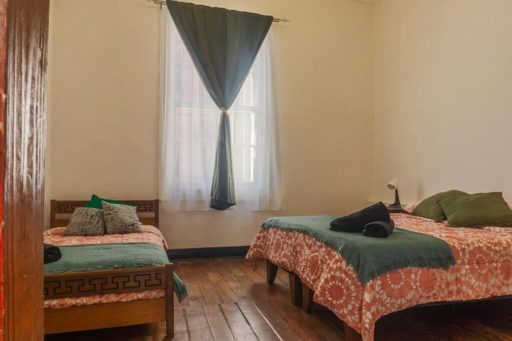 Una habitación con dos camas de madera, una silla, un ventanal con cortinas traslúcidas y una lámpara fijada a la pared.