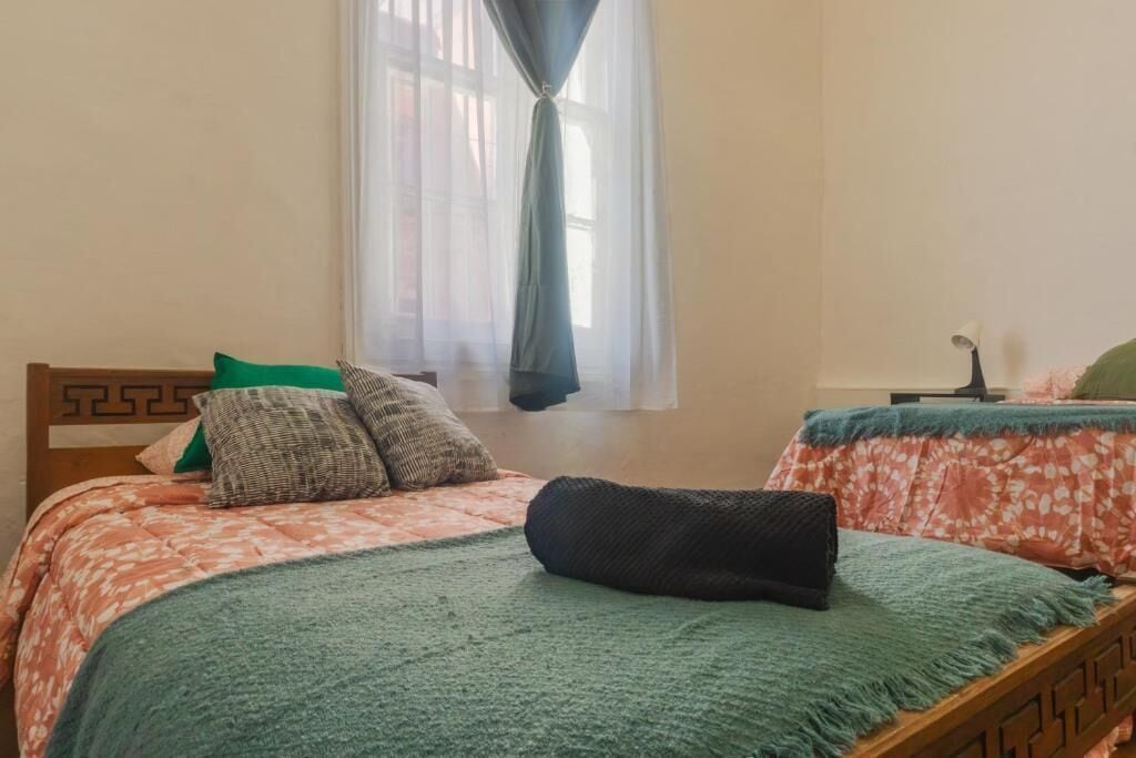 Une chambre avec deux lits, une fenêtre avec des rideaux légers et une petite table avec une lampe.