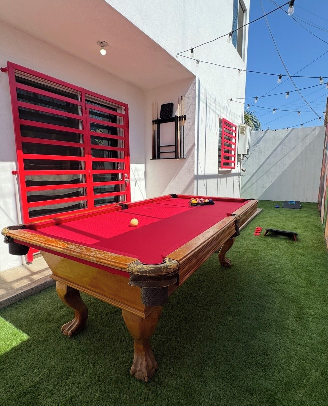 Une table de billard recouverte d’un drap de feutre rouge, entourée d’un gazon artificiel vert, adossée à un mur blanc orné de fenêtres à cadre rouge.