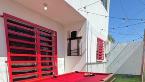 Une table de billard recouverte d’un drap de feutre rouge, entourée d’un gazon artificiel vert, adossée à un mur blanc orné de fenêtres à cadre rouge.