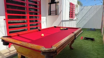 Une table de billard recouverte d’un drap de feutre rouge, entourée d’un gazon artificiel vert, adossée à un mur blanc orné de fenêtres à cadre rouge.
