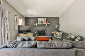 A living room with a grey sofa, a television, a fireplace, and a shelf with decorative items. - Delightful 6-bedroom house in charming Hensonville perfect for families (Hensonville)
