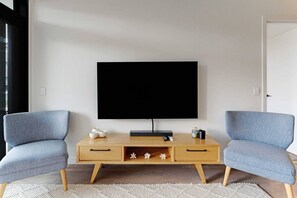 A modern living room with a flat-screen TV mounted on a wooden console table, flanked by two matching armchairs. - The Eden Terrace Retreat with Parking and Balcony (Auckland Central)
