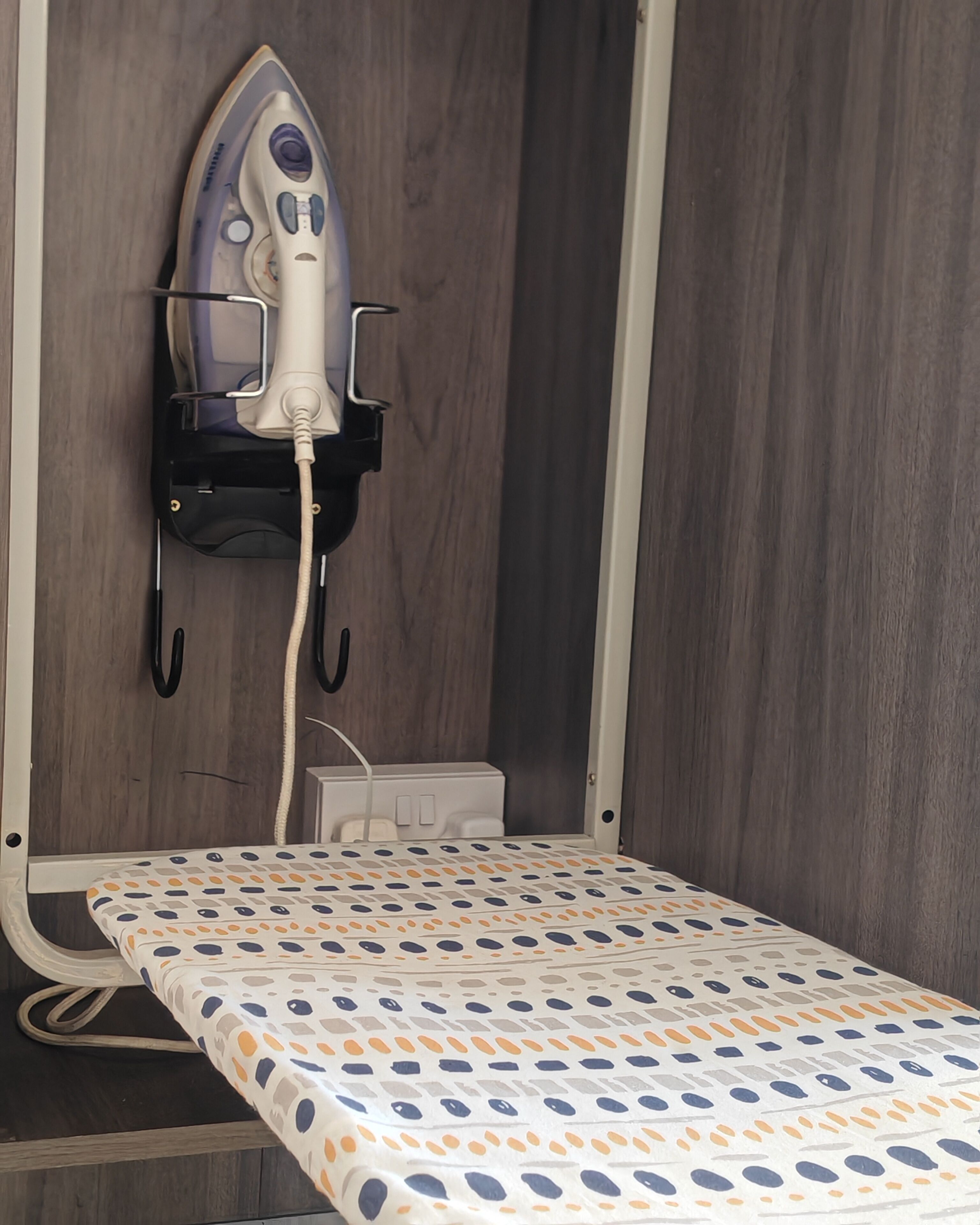 A compact bed with a patterned bedspread, a wall-mounted iron, and a power outlet.