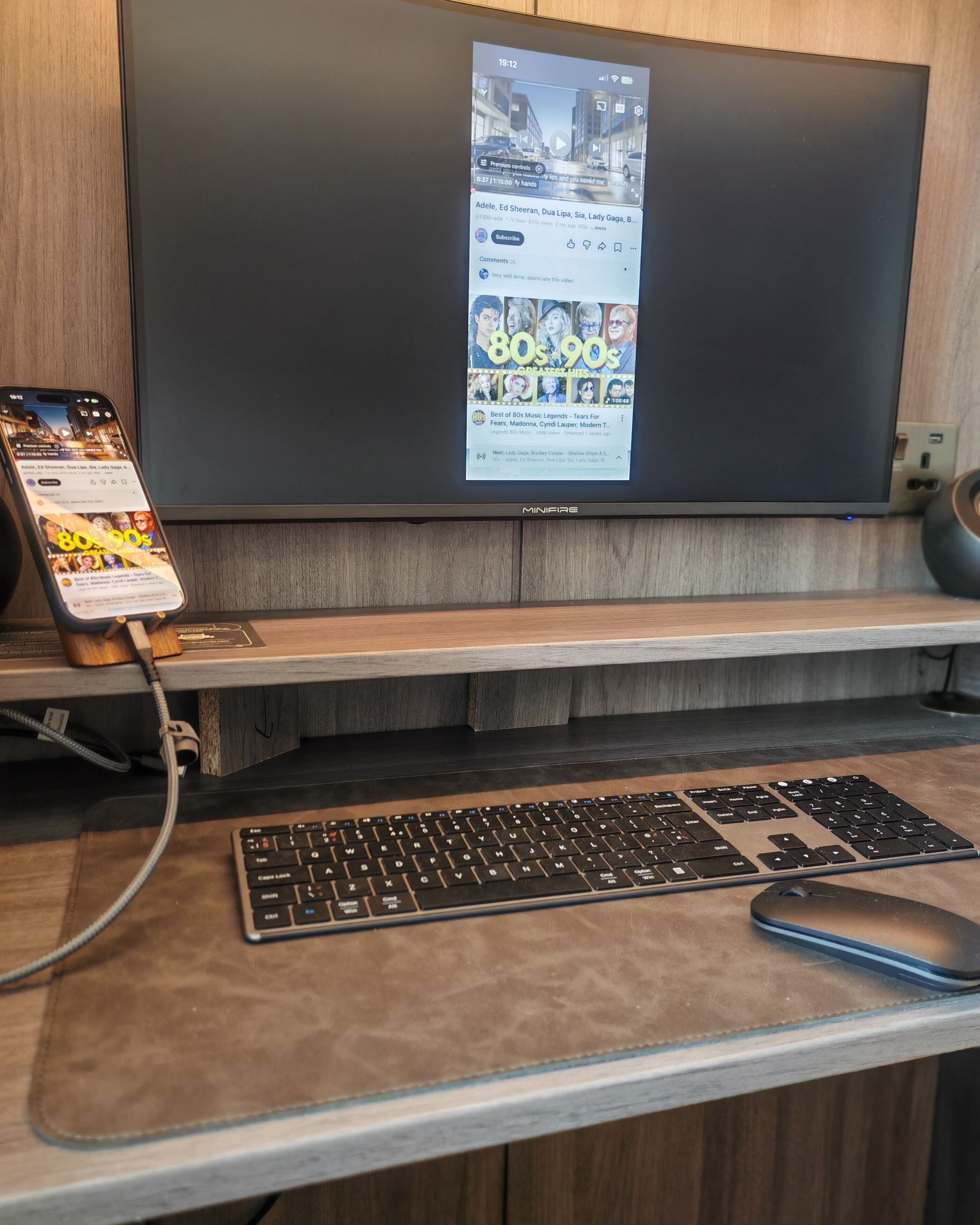 A desk with a laptop, keyboard, and mouse, a smartphone charging, and a television displaying a music video.