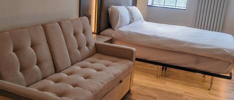 Un dormitorio moderno con sofá, cama, piso de madera y ventana con persianas.