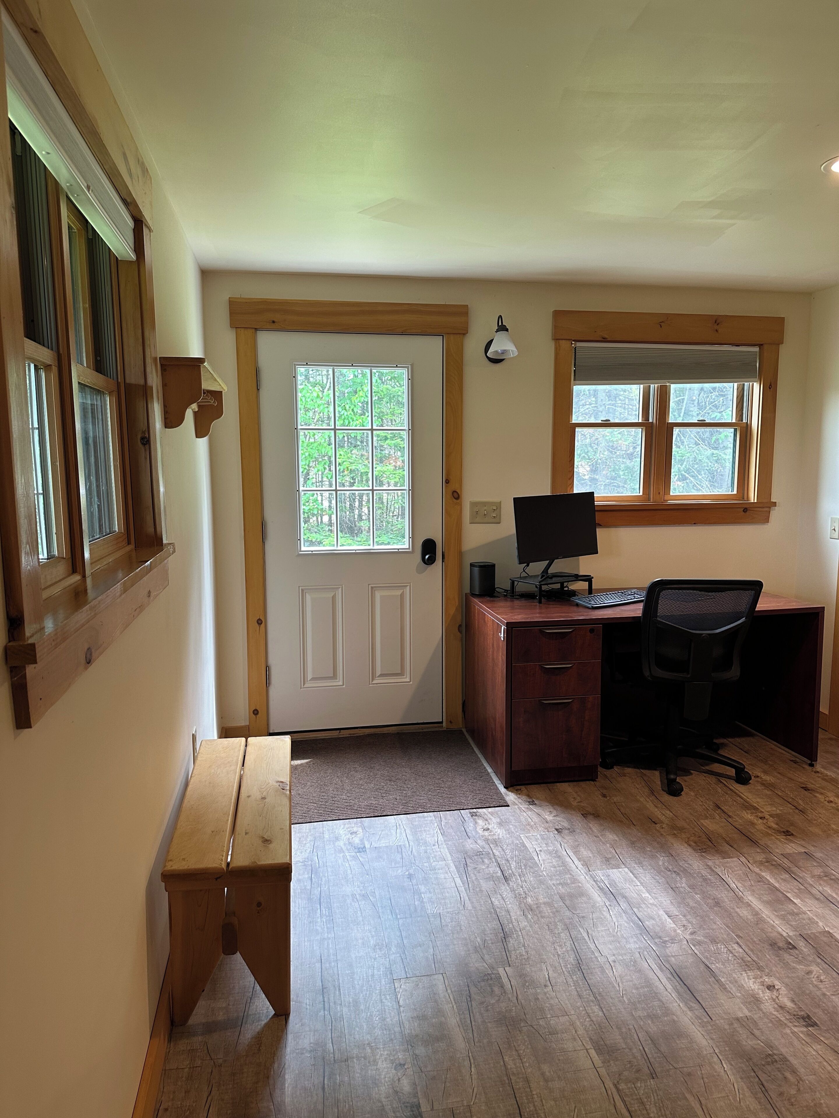 A room with a wooden desk, a computer, and a chair. There is a door with a window, and a bench is visible in the corner.
