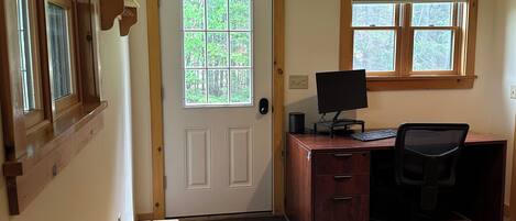 A room with a wooden desk, a computer, and a chair. There is a door with a window, and a bench is visible in the corner.