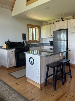 A kitchen with a central island, a stainless steel refrigerator, wooden cabinets, and a window. - Rangeley Sunset Cabin - Lake & Snowmobile Access (Rangeley)