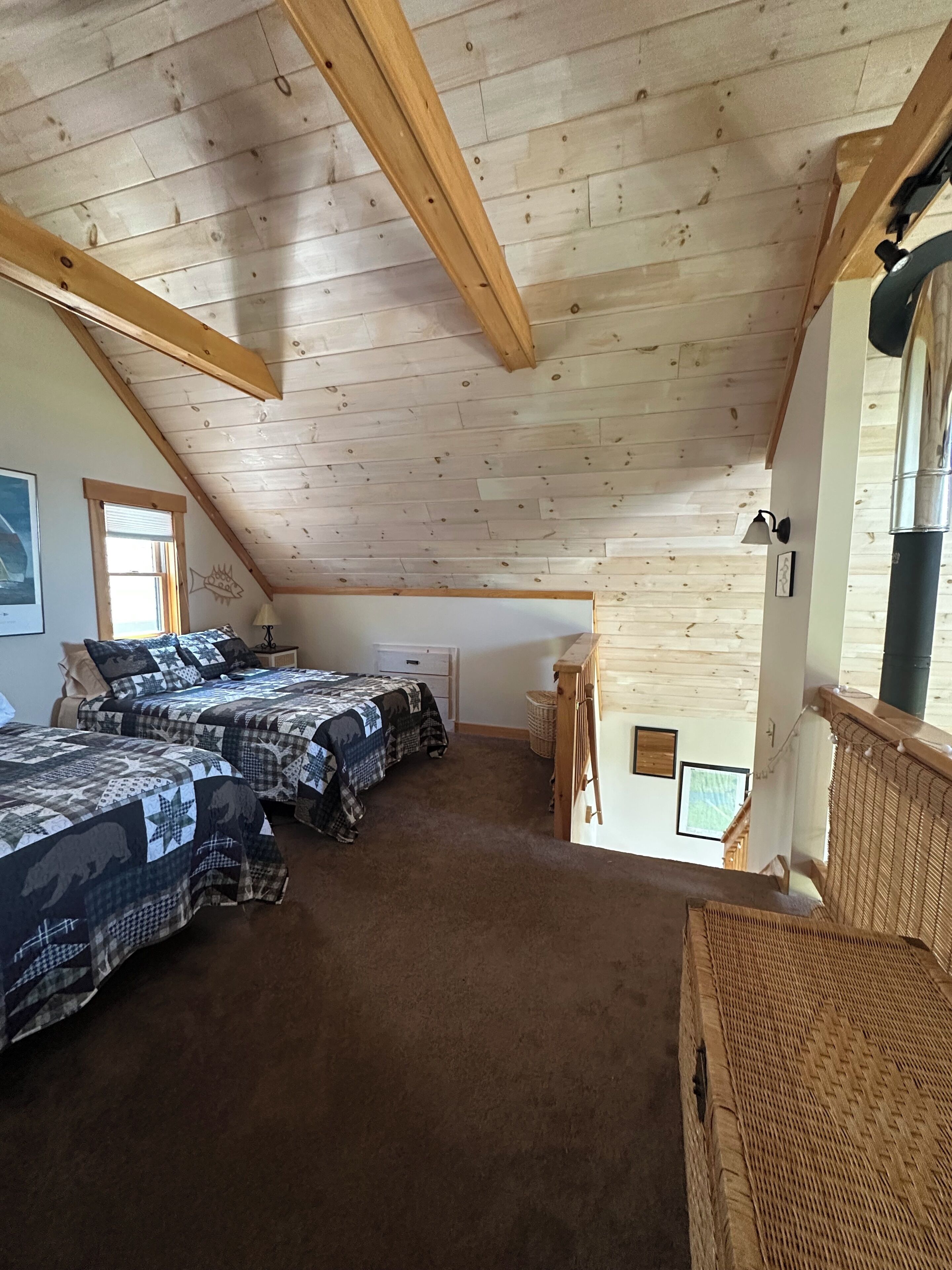 A room with a sloped ceiling, wooden beams, two beds with patterned bedding, a small table, and a wicker chair.