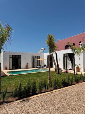 Ein modernes Haus mit Swimmingpool, einer überdachten Terrasse und einem Gebäude mit rotem Dach.