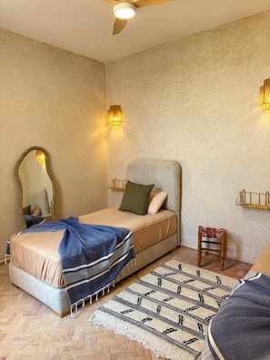 Ein Schlafzimmer mit einem Bett, einem Spiegel, einem Deckenventilator, Wandleuchten, einem kleinen Hocker und einem gemusterten Teppich.