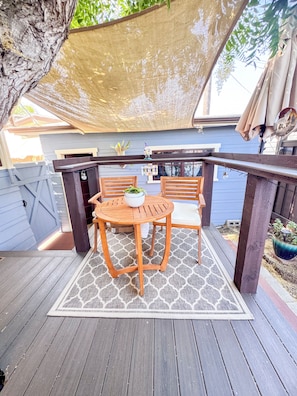 Un terrazzo in legno con tavolo e sedie, un pergolato sopra e una casa blu sullo sfondo.