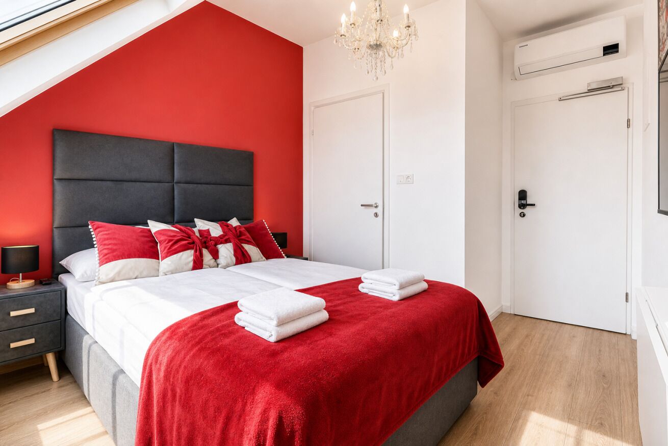Une chambre moderne avec un mur d’accent rouge, un grand lit avec une literie blanche et des oreillers rouges, une table de chevet avec une lampe et un lustre.