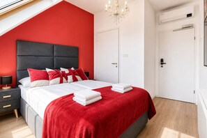 A modern bedroom with a red accent wall, a large bed with white bedding and red pillows, a nightstand with a lamp, and a chandelier. - Bright AC Room Near Hauptbahnhof (Vienna)