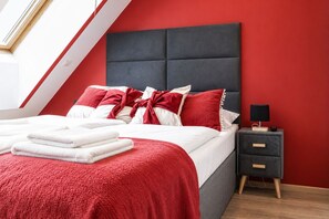 A modern bedroom with a red wall, a grey upholstered headboard, a bedside table with a lamp, and neatly folded white towels. - Bright AC Room Near Hauptbahnhof (Vienna)
