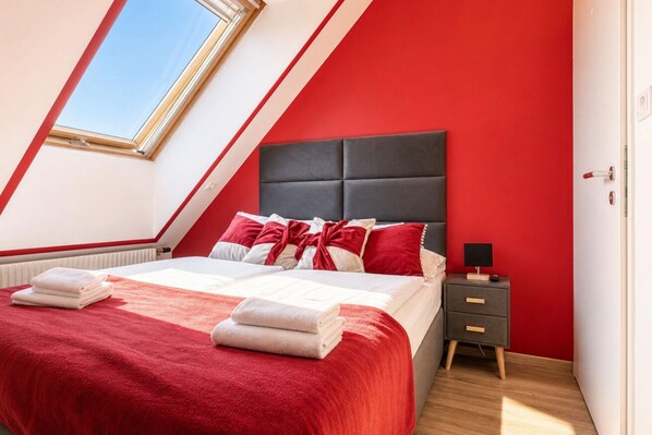 A bedroom with a sloped ceiling, a bed with red and white bedding, a nightstand with a lamp, and a window with a view.